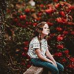 Serene girl sitting on park bench among vibrant red flowers, peaceful outdoor scene, reflective mood, nature tranquility, beautiful floral backdrop. Bright, natural light highlighting her thoughtful expression in a peaceful garden.